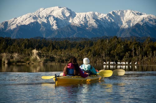 The scene depicts two people paddling a yellow tandem kayak across the calm surface of a lake.