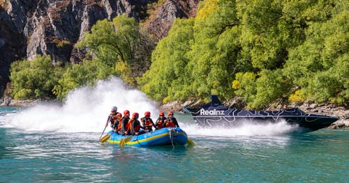 The image shows a group of people aboard an inflatable raft, alongside a black jet boat speeding past.