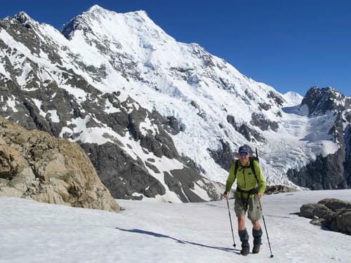 The hiker in the frame is walking across a snowy mountain pass, with New Zealand's highest peak—Aoraki / Mount Cook—in the background.