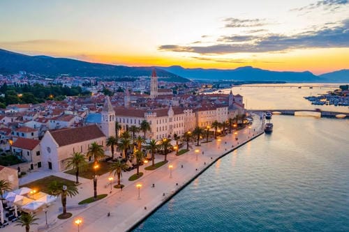 Aerial view of a historic coastal town with palm-lined promenade, lit street lamps, and buildings glowing at dusk beside a calm harbor.