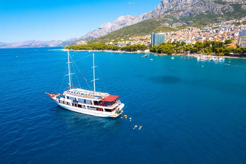 A large white tourist sailboat floats in bright blue waters, with people swimming nearby and a coastal town backed by mountains in the background.