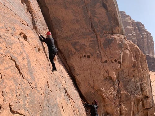 A climber in black gear and a red helmet scales a desert rock face, while a partner belays from below.