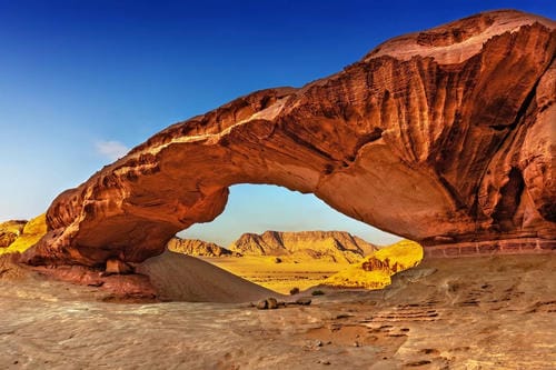 A massive natural sandstone arch dominates the desert landscape, framed by distant rock formations under a clear blue sky.