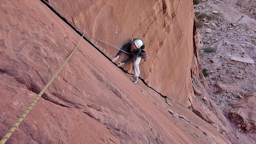 A climber in a white helmet ascends a steep sandstone crack, secured by a rope anchored to the rock above.