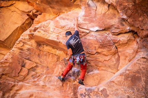 A climber in a black shirt and red pants grips handholds on a textured, sunlit sandstone wall.