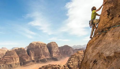A climber in a bright green shirt ascends a red sandstone cliff, with a vast desert canyon and layered rock formations in the background.