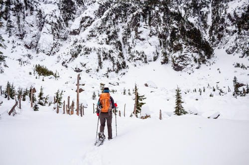 A snowshoer with an orange backpack treks through deep snow toward a snow-covered mountain slope.