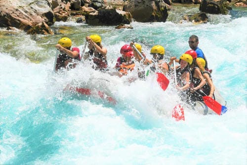 A raft plows through frothy rapids, sending large splashes of water over the bow.