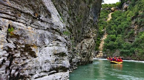 A raft drifts down a calm, green river lined by steep, rocky canyon walls.