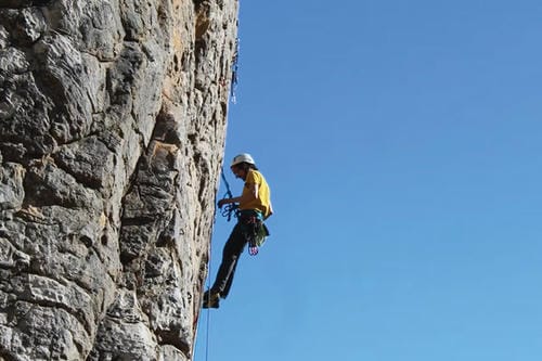 A climber in a yellow shirt and white helmet ascends a vertical, textured grey rock face under a clear blue sky, using ropes and climbing equipment.