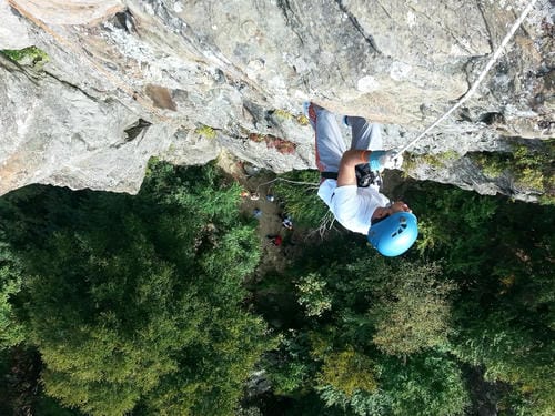 A climber in a blue helmet rappels down a steep, grey rocky cliff, with lush green trees visible far below.