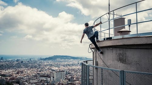 A man in a grey t-shirt and dark pants perches on the edge of a tall building, overlooking a vast cityscape and the sea under a partly cloudy sky.