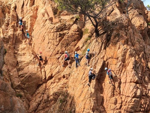 A group of climbers in helmets and harnesses ascend a rugged, reddish-brown rock face equipped with safety ropes and gear.