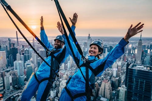 Two people in blue jumpsuits and helmets celebrate with raised arms while attached to a city-high safety harness, with a stunning sunset over a sprawling urban skyline in the background.