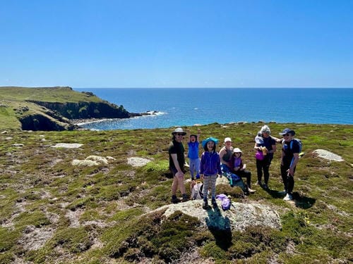 A group of people pose for a photo on a grassy coastal cliff with the bright blue sea and a clear sky in the background.