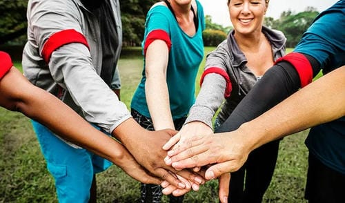 A diverse group of people stack their hands together in a show of teamwork and unity in a grassy park.