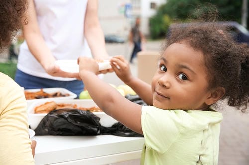 A young girl happily receives a meal from a volunteer at an outdoor food distribution event.