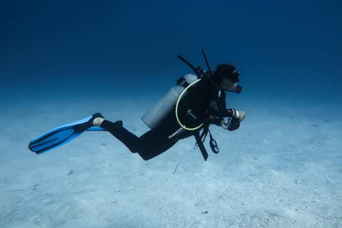 A scuba diver in full gear swims horizontally over a sandy ocean floor in clear blue water.