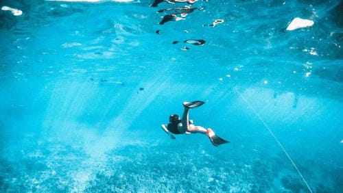 A snorkeler with fins swims underwater in bright turquoise water, with other swimmers visible near the surface above.