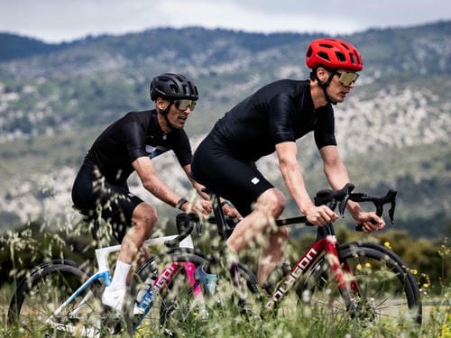 Two male cyclists, one with a black helmet and the other with a red helmet, ride closely together through a grassy, mountainous area.