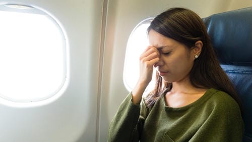 A woman appears uncomfortable, rubbing her eyes while seated in an airplane, likely experiencing motion sickness.