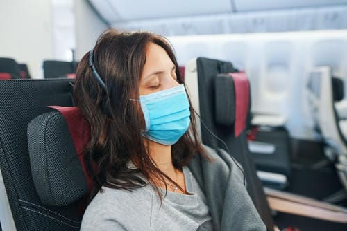 A woman rests with her eyes closed on an airplane, wearing a mask and headphones, possibly trying to ease travel discomfort.