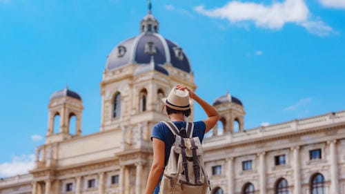 A tourist wearing a hat and backpack stands in front of the Kunsthistorisches Museum in Vienna, Austria.