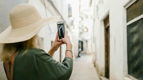 A tourist in a wide-brimmed hat takes a photo of a narrow street with her smartphone.
