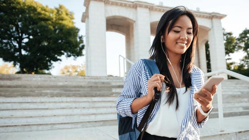 A young woman wearing earphones and carrying a backpack walks while using her smartphone outdoors.