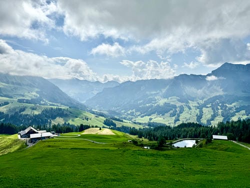 A panoramic view of rolling green pastures, traditional alpine buildings, and distant misty mountains under a partly cloudy sky.