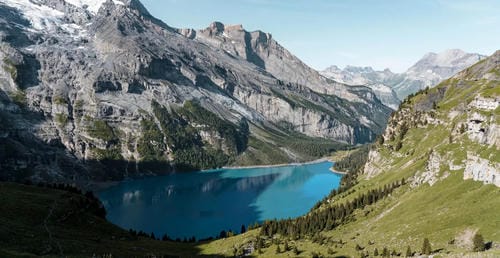 A turquoise alpine lake nestles in a rugged mountain valley surrounded by steep, snow-capped peaks and lush green slopes.