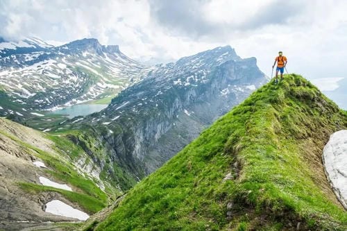 A hiker stands triumphantly on a grassy mountain ridge, overlooking a vast landscape of snow-capped peaks and a distant glacial lake.