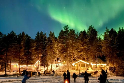 Green northern lights arc across the sky above a snow-covered village of lit wooden cabins and pine trees, as people admire the view.
