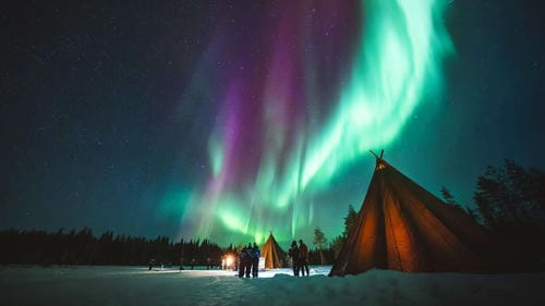 A group of people gather outside lit Sami-style tents beneath a dazzling display of green and purple aurora borealis in a snowy northern landscape.