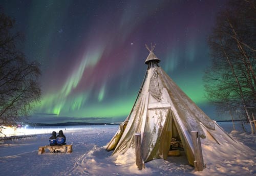 Two people sit on a wooden bench beside a traditional Sami tent, watching the vivid aurora borealis light up the night sky over a frozen lake.