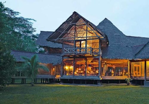 A multi-level thatched lodge in the rainforest glows warmly at dusk, with open-sided communal spaces.