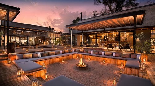 A luxury safari lodge's outdoor fire pit area glows with lantern light under a sunset sky.