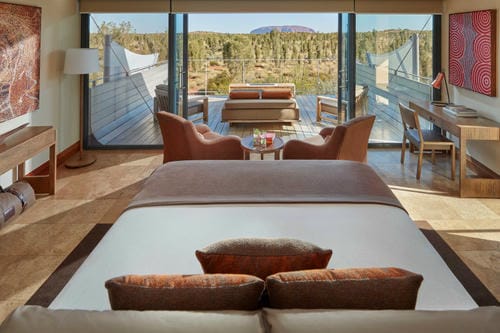 A modern hotel room with floor-to-ceiling windows frames a view of Uluru (Ayers Rock) in the Australian outback.