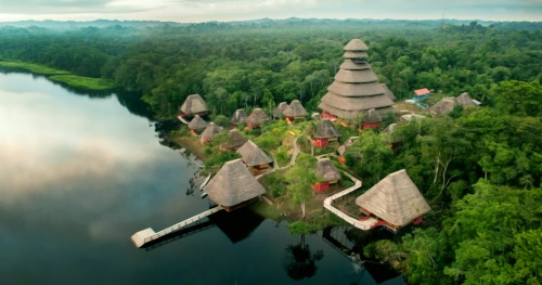 Aerial view of an eco-lodge with thatched-roof huts nestled along a calm river in the Amazon rainforest.