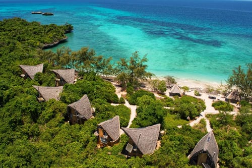 Thatched-roof bungalows sit among lush greenery on a tropical island overlooking turquoise ocean waters.