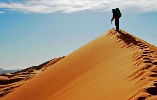 A lone hiker with a backpack and walking stick stands at the sharp crest of a towering sand dune.
