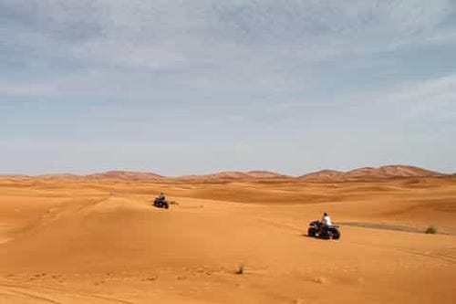 Two people ride quad bikes across a wide, open desert landscape under a pale sky.