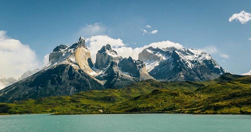A wide view of snow-dusted granite mountains rising above a turquoise lake and rolling green grasslands under a bright sky.