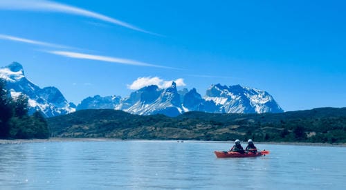 Two people paddle a red kayak across calm blue water, with dramatic snow-capped mountains and a clear sky in the background.