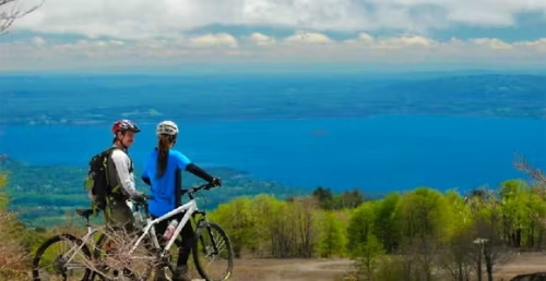 Two mountain bikers pause at a scenic overlook with their bikes, taking in the view of a vast blue lake and distant green hills under a partly cloudy sky.