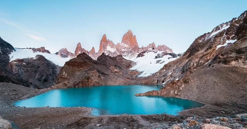 Towering, jagged snow-capped peaks rise above a bright turquoise glacial lake set in a rocky mountain valley.