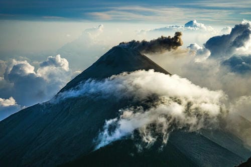 A perfectly conical volcano emits a dark plume of ash, partially surrounded by clouds at sunrise or sunset.