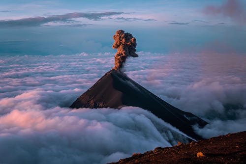 A volcano erupts above a sea of clouds, sending a mushroom-shaped ash cloud into the twilight sky.