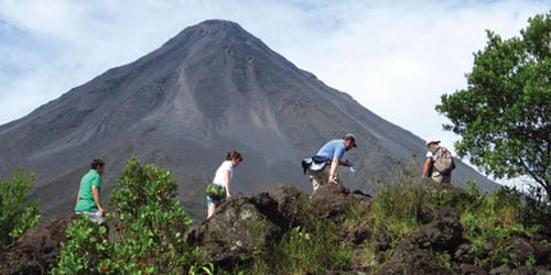 A group of hikers climbs rocky terrain with a steep, symmetrical volcanic cone rising behind them.