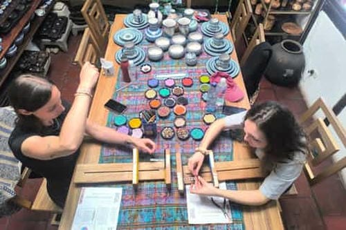 Two women work side-by-side at a table filled with colorful paints, pottery, and art supplies in a craft workshop.